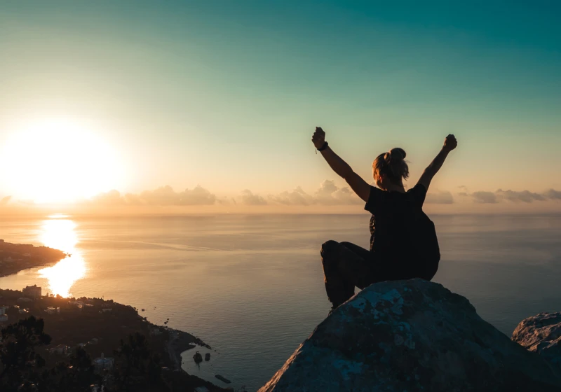 Girl sitting on top of mountain with arms up in celebration watching sunset